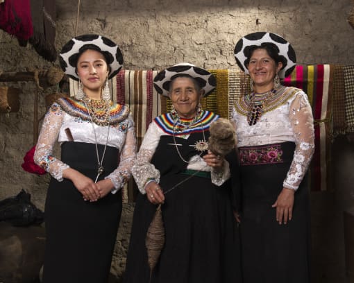 Three generations of women from Saraguro, Ecuador in their traditional costumes that include heavy woolen hats in black and white, bright white blouses, dark black skirts, and colorful jewelry and belts