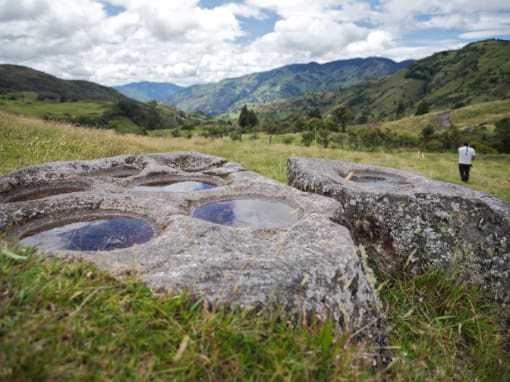 Rocas ancestrales con agujeros tallados, posiblemente utilizados para molienda, se encuentran en medio de un pastizal rodeado de montañas, mientras un hombre camina a lo lejos.